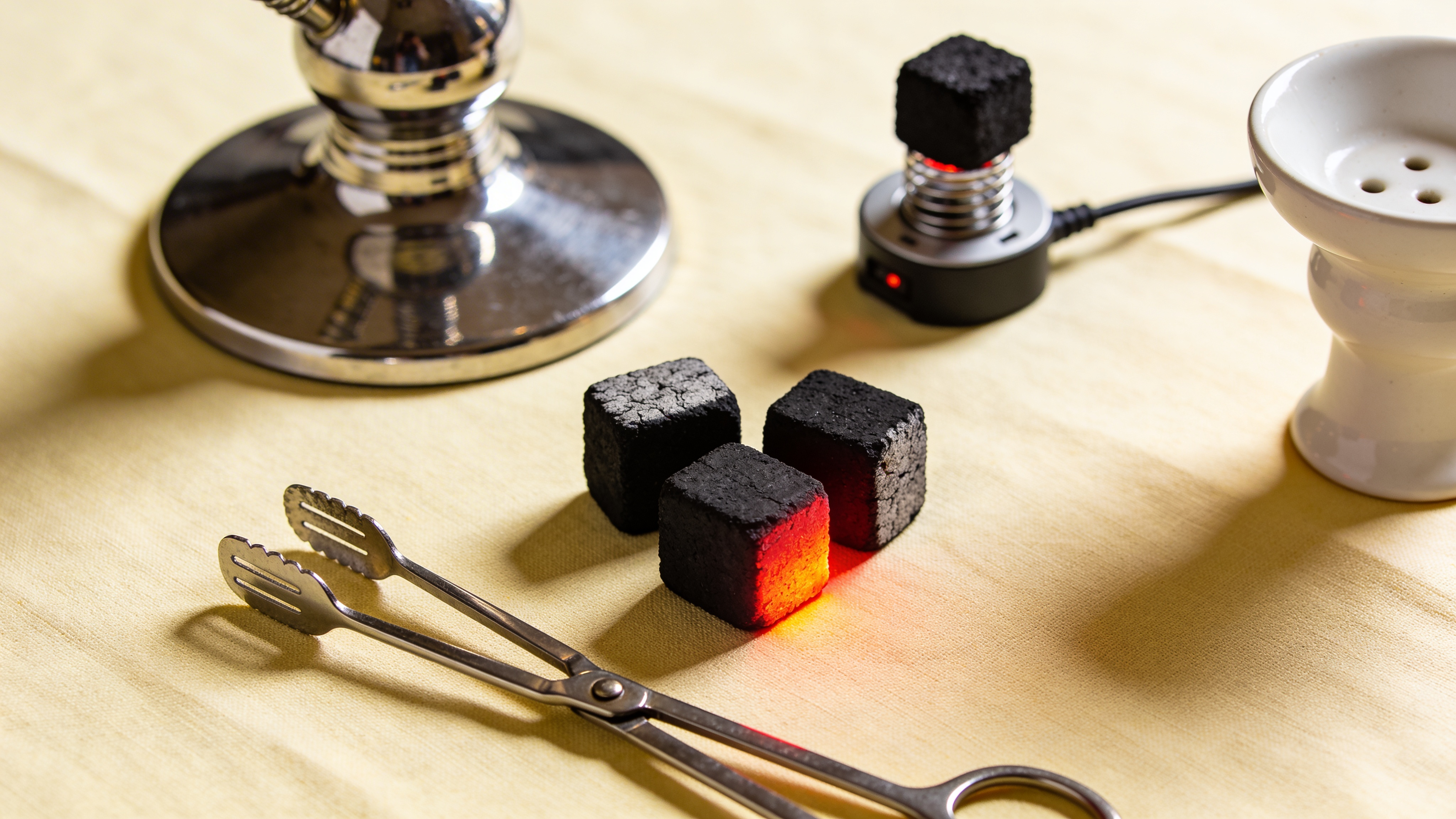 Natural coconut charcoal cubes glowing red-orange beside stainless steel tongs and an electric coal burner — essential hookah charcoal gear on a warm cream surface