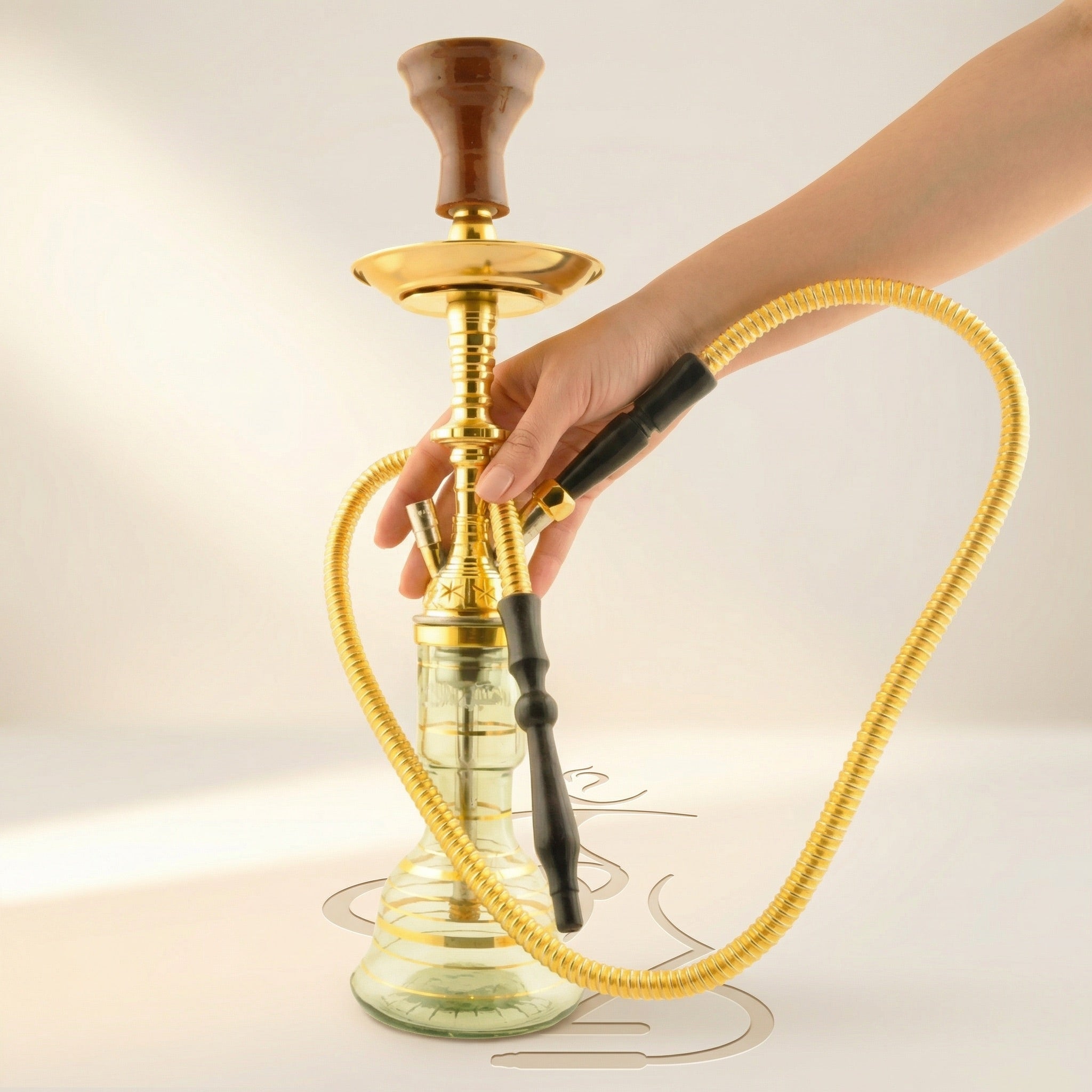 Hand resting on the gold-plated stem hub of the Khalil Mamoon Mini Beast Gold hookah, showing the compact 52 cm Egyptian pipe with its gold coil hose curved around the clear glass base.