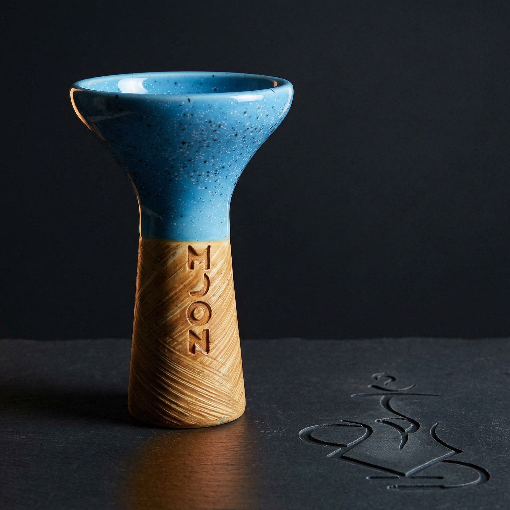 Moon Phunnel blue speckled glazed clay shisha bowl shot in dramatic dark studio lighting on slate, the glaze and hand-stamped MOON wordmark glowing under a single warm key light.