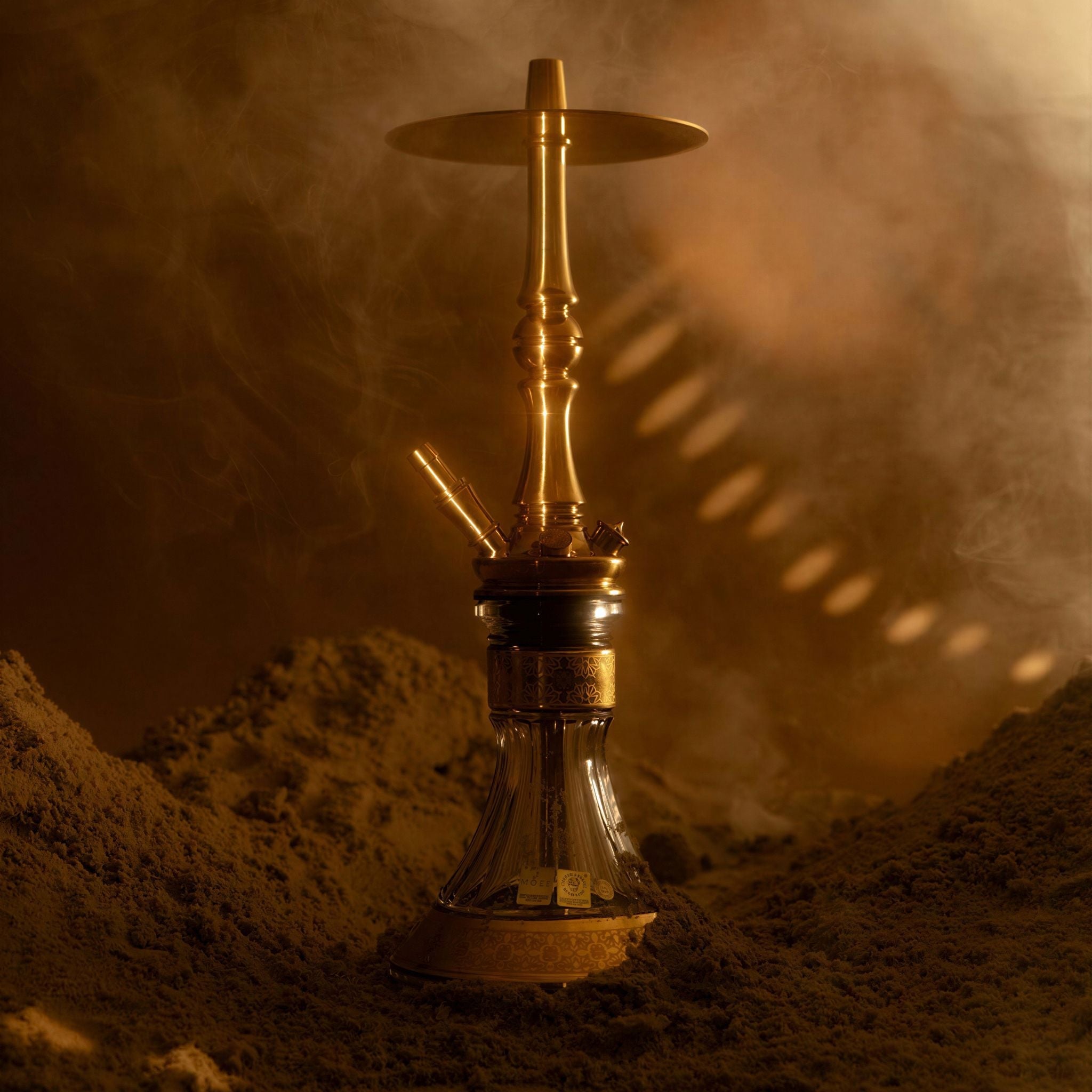 Moze Tradi M Gold traditional shisha in a warm smoky desert setting, polished gold stem on a cut-glass base