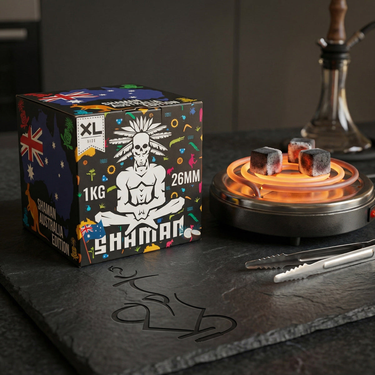 Shaman 26mm coconut charcoal 1kg box on a dark slate countertop beside an electric burner with glowing coals, tongs, and a hookah in the background., image 3 of 15
